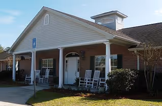 Single-story brick assisted living building entrance with a covered porch, white columns and several rocking chairs.
