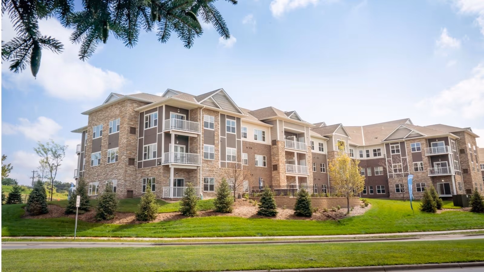 Exterior view of a large, modern senior living facility building with multiple floors, balconies, and a combination of stone and siding on the facade. The building is surrounded by well-maintained green lawns, small trees, and shrubs under a partly cloudy sky.
