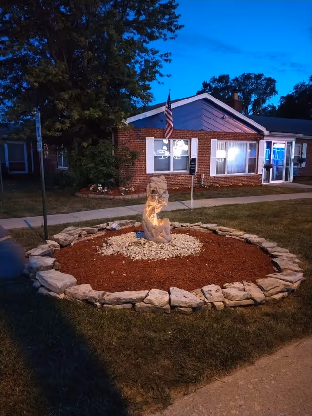 A brick single-story senior living building at dusk with a circular stone-bordered garden and an illuminated rock fountain in the foreground and an American flag by the entrance.
