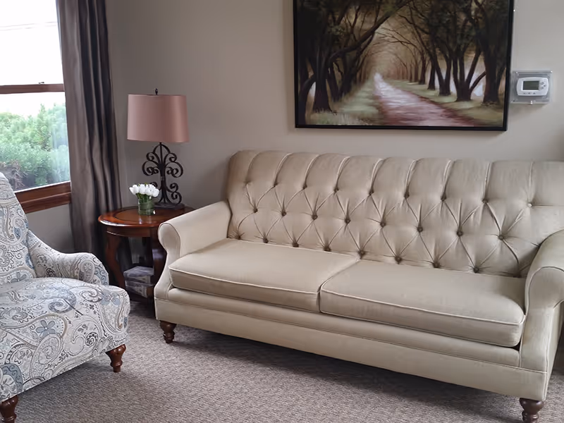 A cozy living room with a tufted beige sofa, patterned armchair, side table with a lamp and flowers, and a framed landscape painting.