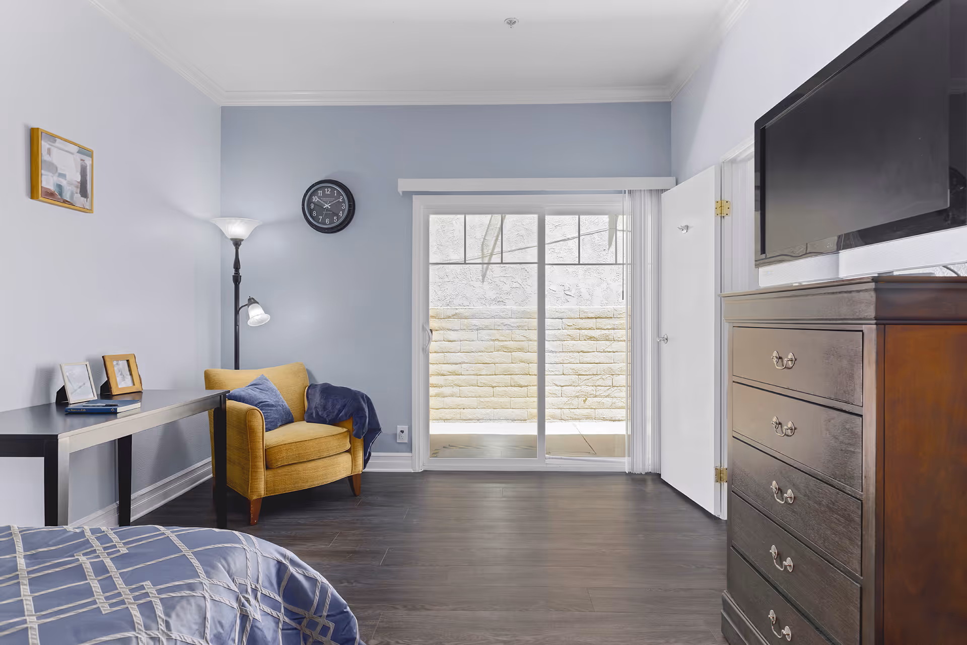 A bright bedroom with a blue and gray color scheme featuring a bed with a patterned comforter, a yellow armchair with a blue throw blanket, a black desk with picture frames and a book, a floor lamp, a wall clock, a sliding glass door leading to an outdoor area, and a dark wooden dresser with a flat-screen TV on top.
