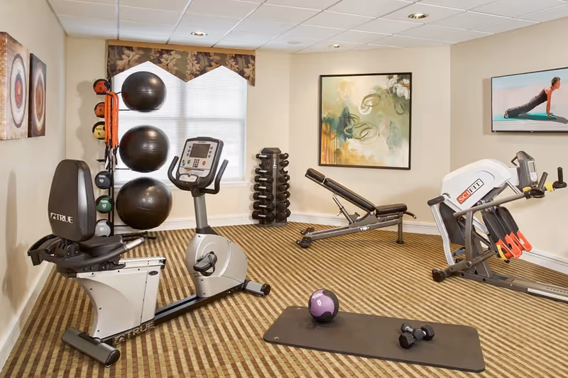 Small indoor fitness room with exercise machines, free weights, stability balls, and a wall-mounted TV.