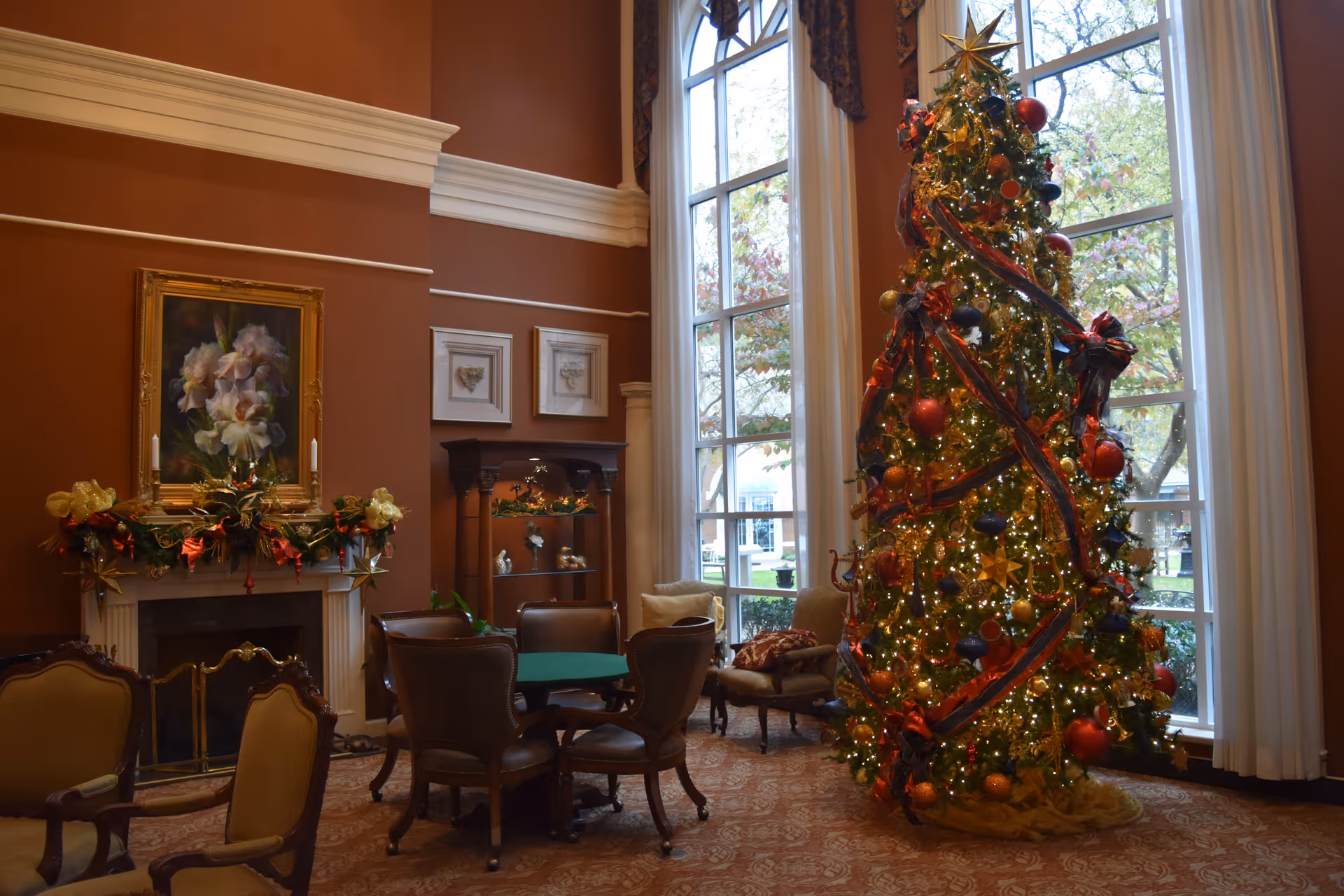 A cozy living room decorated for Christmas with a large, decorated Christmas tree adorned with lights, ribbons, and ornaments near tall windows. The room features a fireplace with festive garland and candles, a painting above the mantel, several upholstered chairs, and a small round table. The walls are painted a warm brown color.