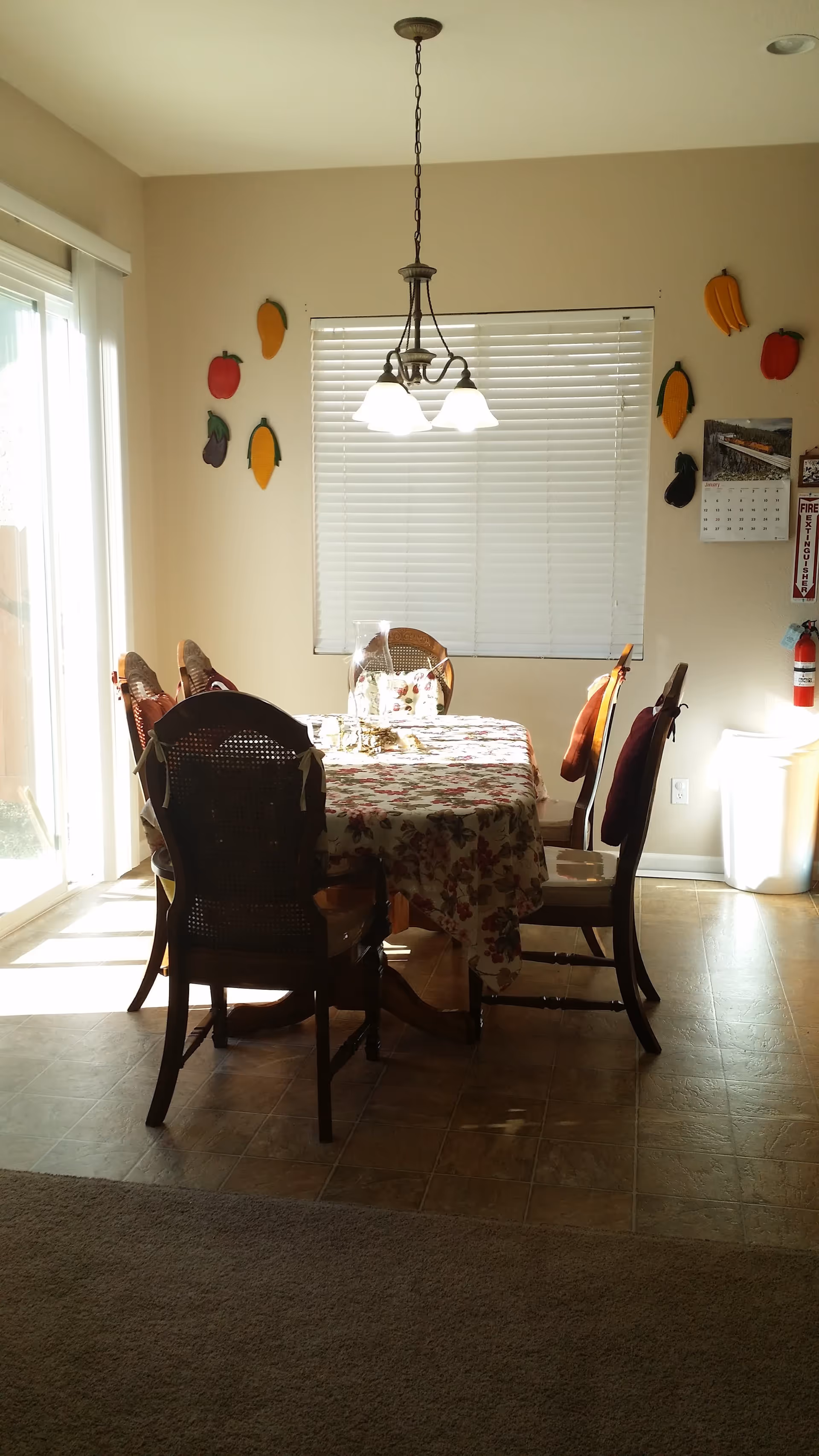 A dining area with a rectangular table covered with a floral tablecloth and surrounded by six wooden chairs with cushions. Above the table hangs a three-light chandelier. The wall behind the table has a window with closed blinds and decorative fruit-shaped wall hangings. To the right, there is a fire extinguisher mounted on the wall and a calendar. Sunlight streams in from a sliding glass door on the left.