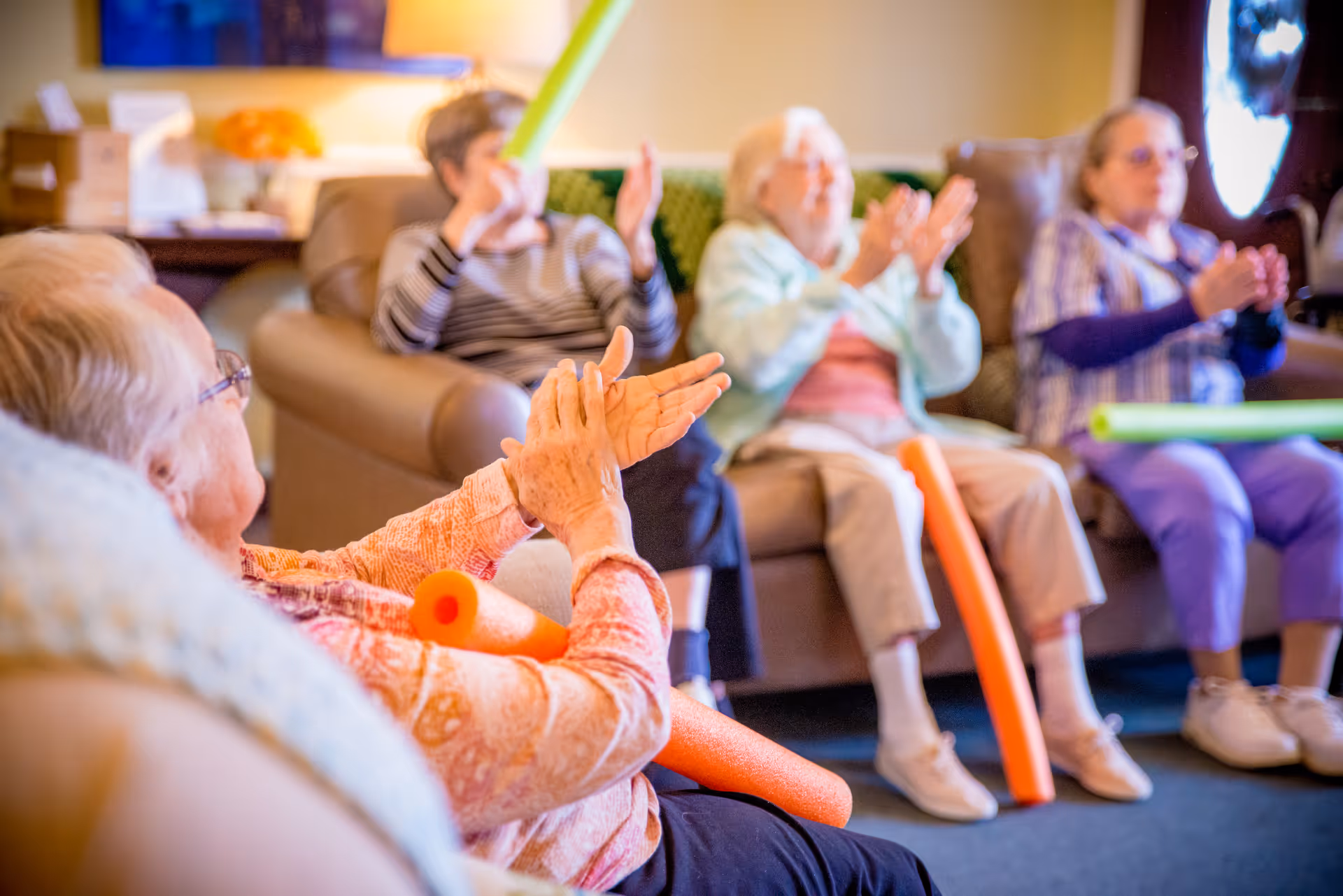 A group of elderly women sitting in a cozy living room, clapping and holding colorful foam noodles, engaging in a group activity.