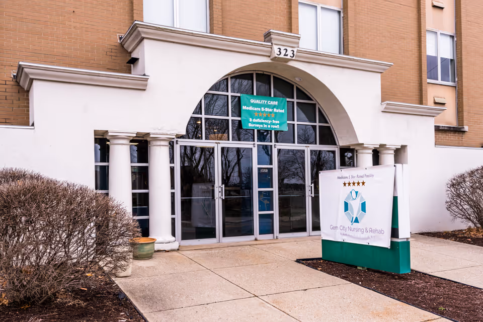 Front entrance of Gem City Nursing and Rehabilitation Center with glass doors, columns, and a freestanding sign displaying the facility name.