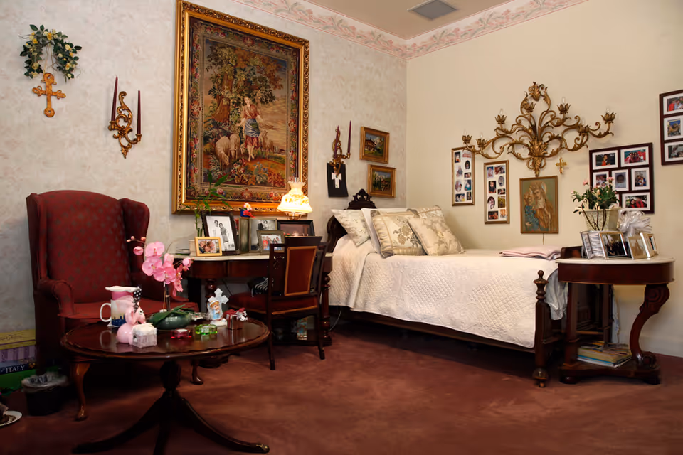 A cozy bedroom with a single bed covered in a white quilt and decorative pillows. The room features a red upholstered armchair, a wooden desk with framed photos and a lamp, and a round wooden table with various items including a pink orchid. The walls are decorated with framed pictures, a large tapestry, and ornate candle holders.