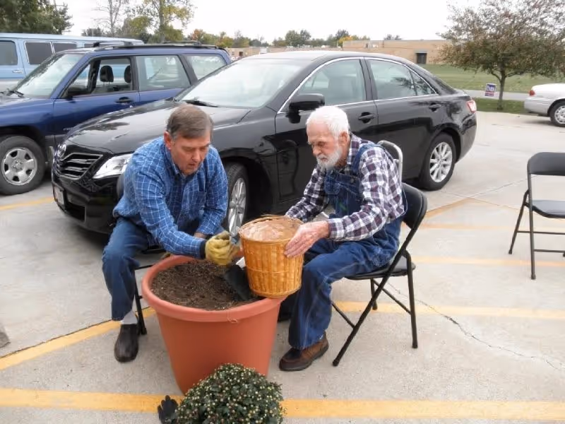 Two elderly men plant soil into a large flower pot in an outdoor parking lot.