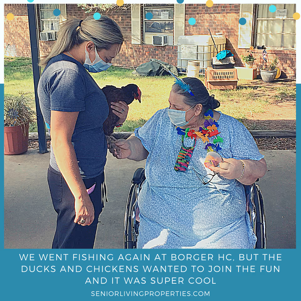 A woman in a wheelchair wearing a blue dress and colorful lei interacts with another woman standing and holding a chicken. Both women are wearing face masks. They are outside near a brick building with potted plants and air conditioning units visible in the background.