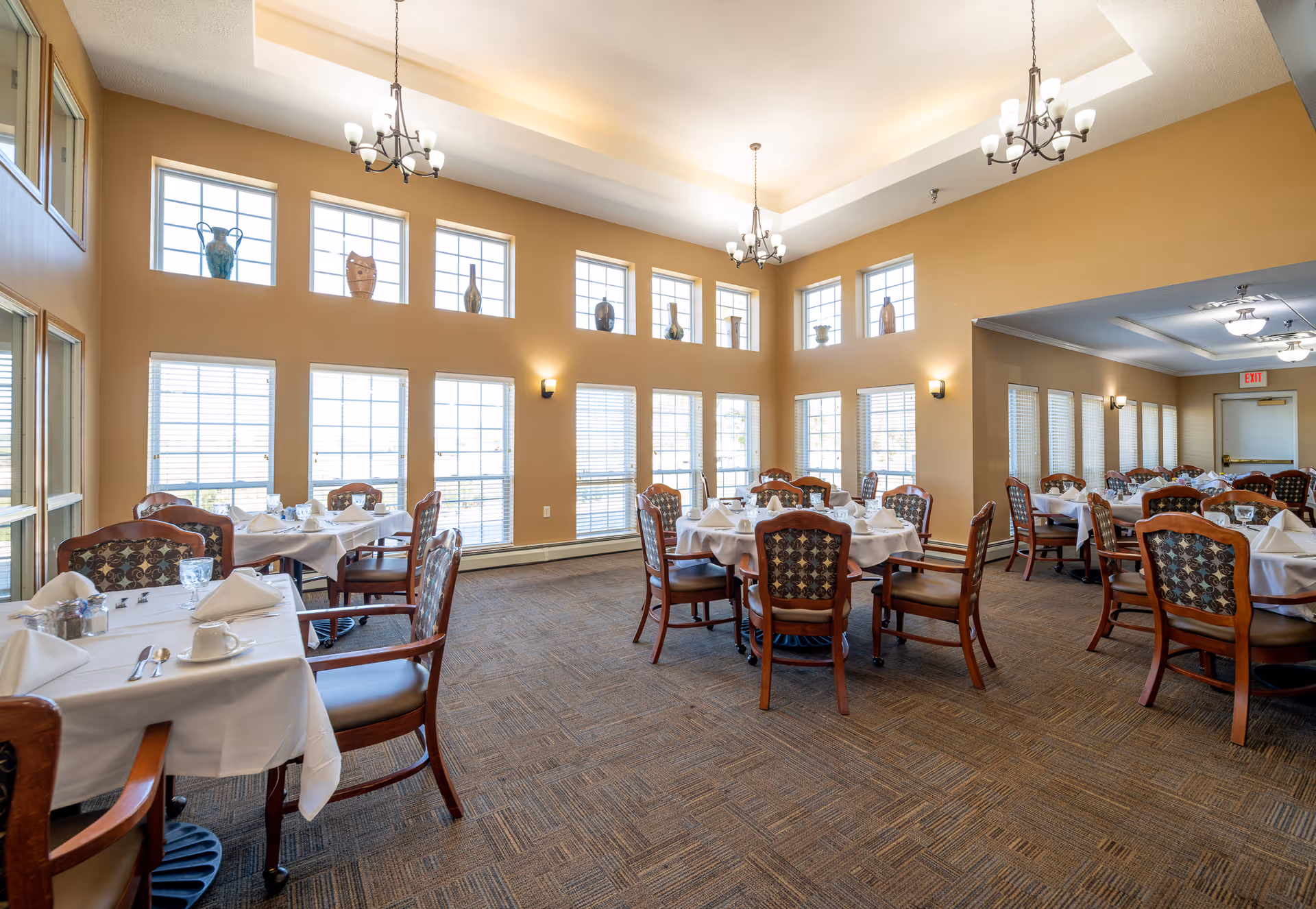 A spacious dining room with multiple round and rectangular tables covered with white tablecloths, set with plates, cups, napkins, and silverware. The room has large windows with blinds allowing natural light to fill the space, beige walls, carpeted floor, and decorative vases displayed on a ledge near the ceiling. Elegant chandeliers hang from the ceiling providing additional lighting.