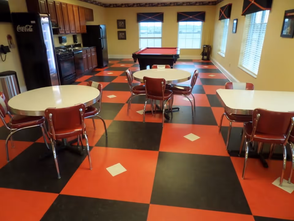 A recreational room with a red and black checkered floor featuring white diamond accents. The room contains several tables with red chairs, a pool table with a red felt top near the windows, and a kitchenette area with a Coca-Cola vending machine, refrigerator, stove, and cabinets. The walls are painted yellow and there are multiple windows with blinds and valances.