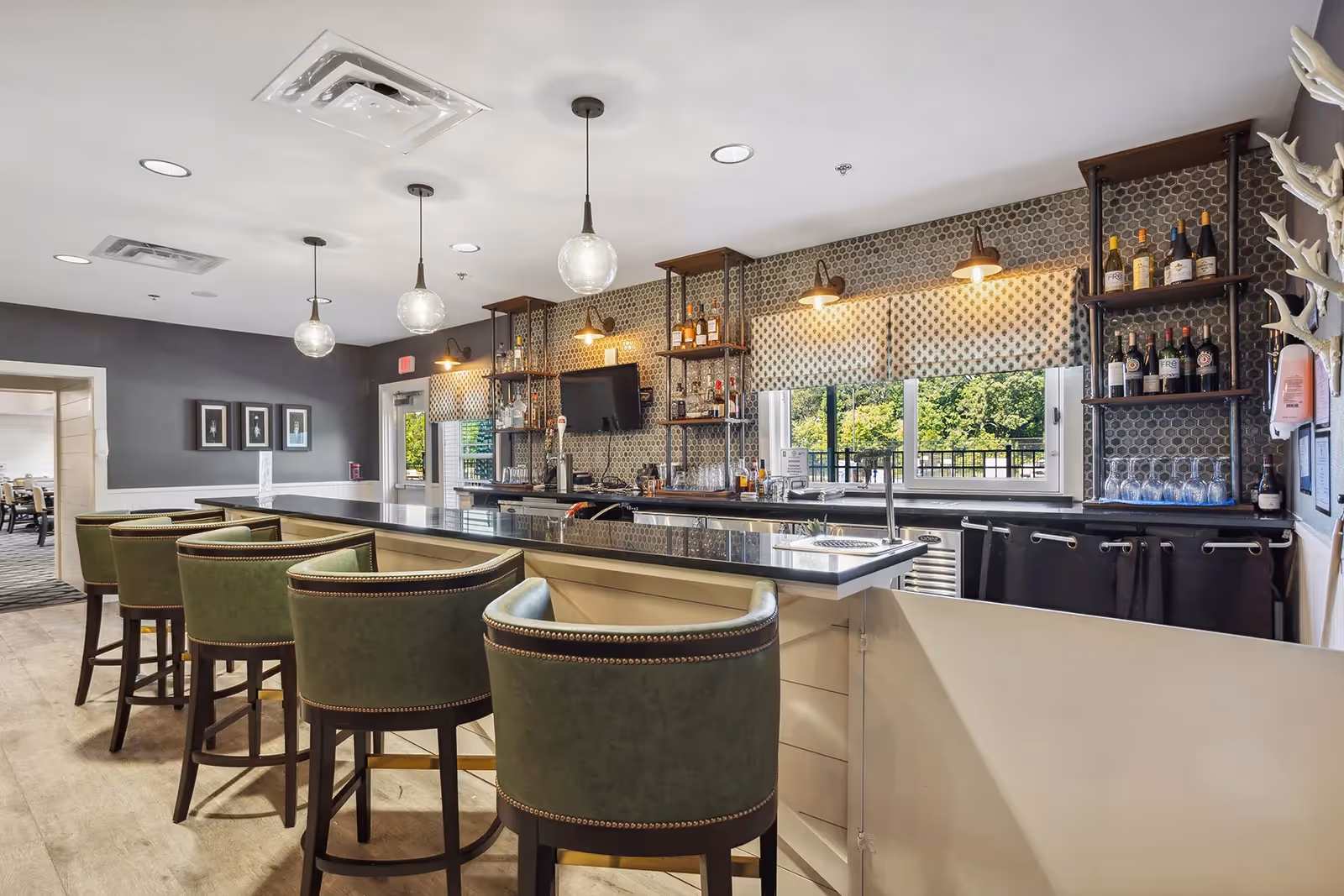 Interior view of a stylish bar area with a long counter, green cushioned bar stools, hanging pendant lights, and shelves stocked with bottles and glasses. Large windows with patterned blinds allow natural light to brighten the space, and a door leads to an adjacent room.