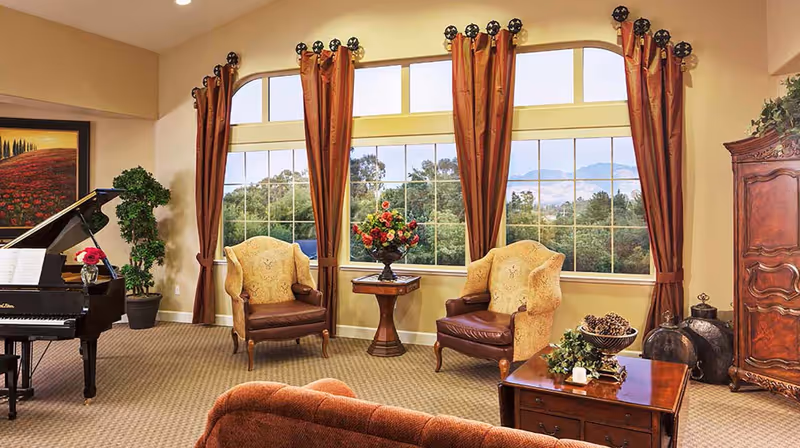 A cozy living room with large windows covered by rust-colored curtains, two upholstered armchairs with wooden legs, a wooden side table with a floral arrangement, a grand piano with sheet music and a flower vase, a wooden coffee table with decorative items, and a large wooden cabinet. The room has a beige carpet and a scenic view of trees and mountains outside the windows.