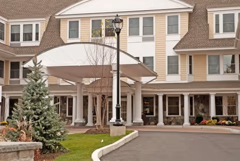 Front exterior view of a multi-story senior living facility with beige siding, white trim, and a covered entrance supported by white columns. There is a black street lamp in front, landscaping with evergreen trees and shrubs, and a paved driveway leading to the entrance.