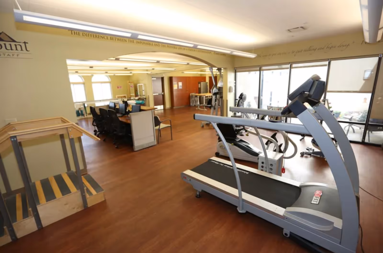 Interior view of a rehabilitation facility room with exercise equipment including a treadmill and stationary bike, a desk with chairs and computers, wooden flooring, large windows letting in natural light, and a small set of stairs with handrails.