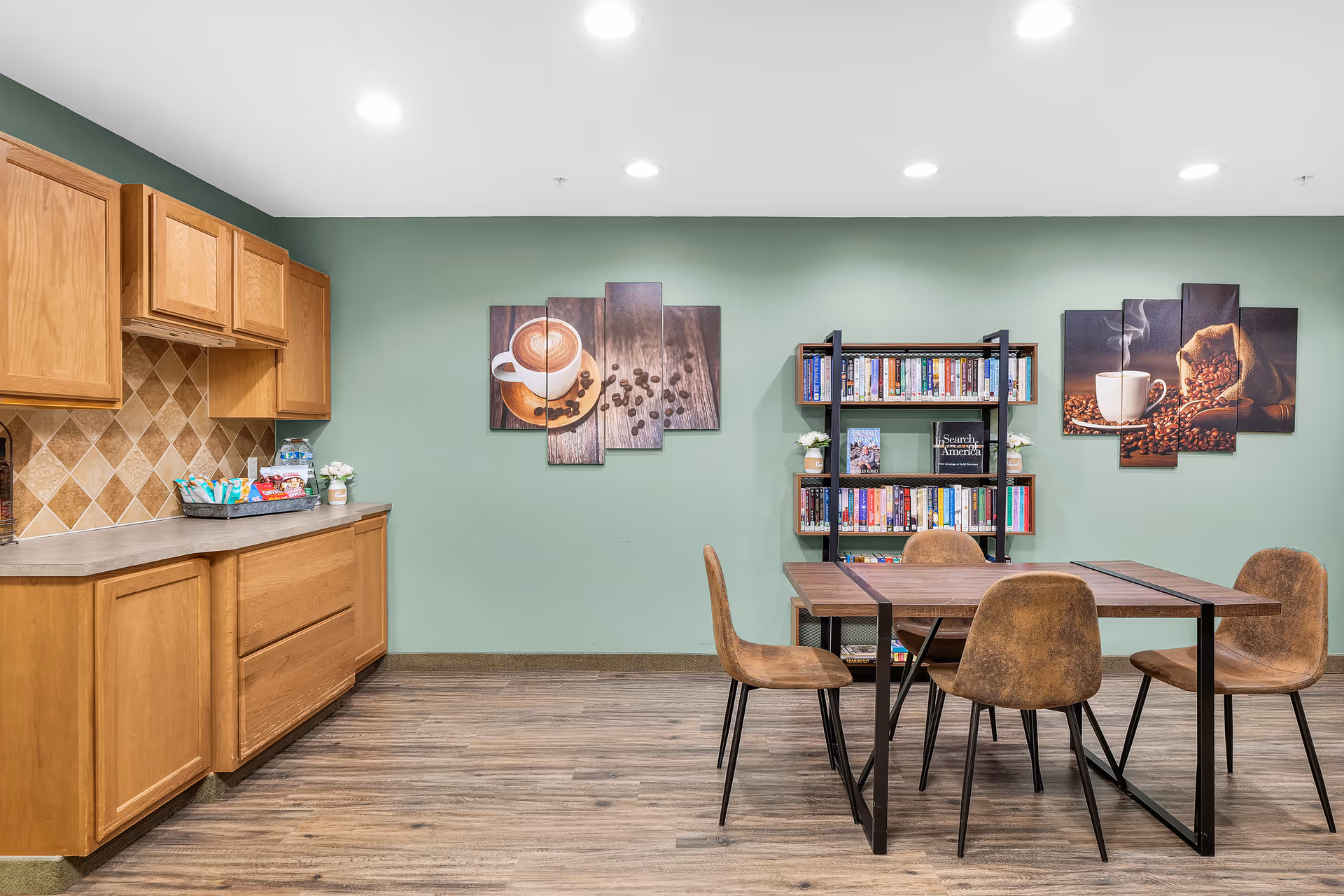 A cozy common area with a wooden table and four brown chairs on a wood floor. On the left side, there is a kitchenette with wooden cabinets and a countertop holding snacks and a small plant. The back wall is painted green and decorated with two multi-panel coffee-themed artworks and a black bookshelf filled with books and small plants.