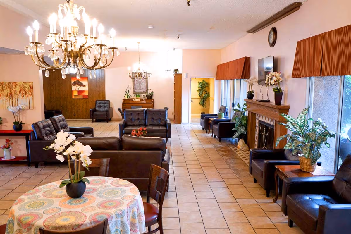 Bright retirement home common lounge with leather sofas and armchairs, chandeliers, a fireplace, and a round table with a floral centerpiece.