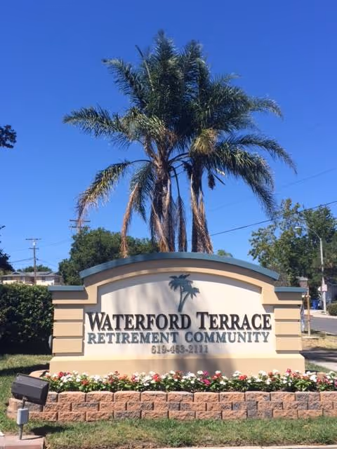 Outdoor sign for Waterford Terrace Retirement Community with a palm tree logo, surrounded by a flower bed and a tall palm tree in the background under a clear blue sky.