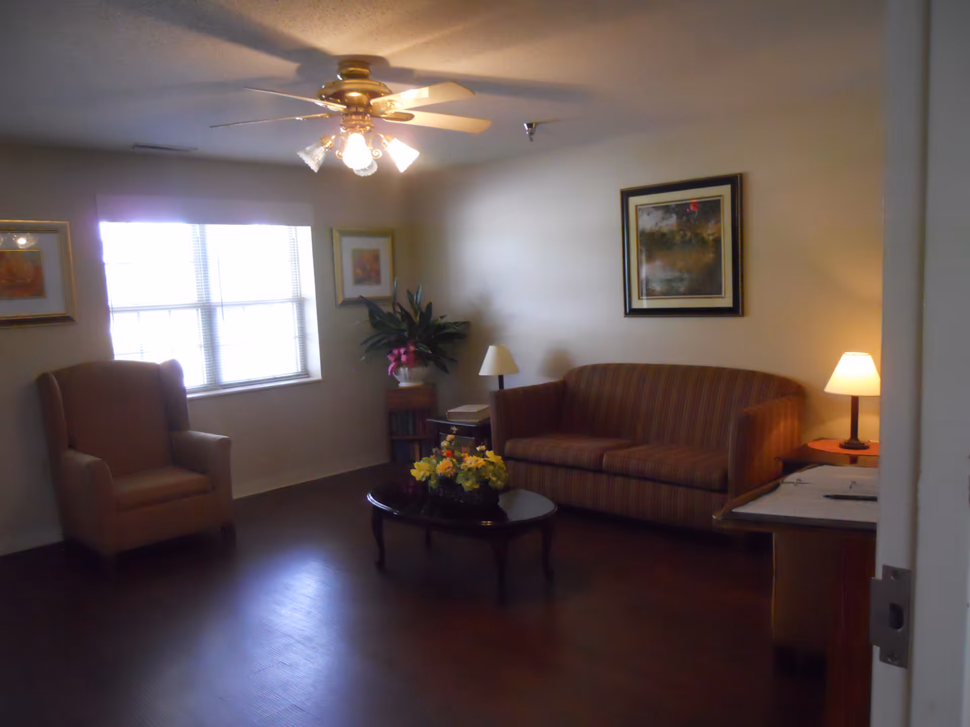 A cozy living room with a striped sofa, an armchair, a coffee table with a flower arrangement, a side table with a lamp, framed artwork on the walls, a ceiling fan with lights, and a window with blinds letting in natural light.