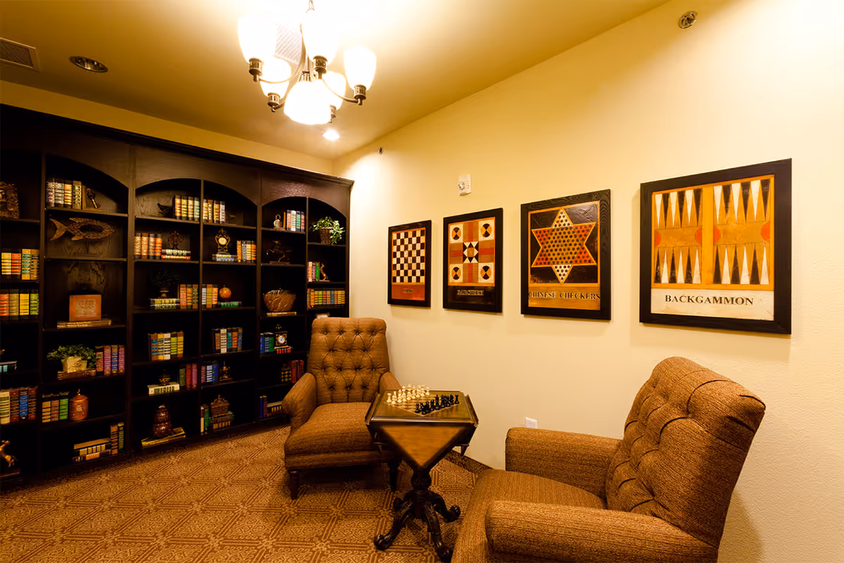 Cozy lounge with two tufted armchairs facing a small table with a chess set, a large bookshelf filled with books along the left wall, and framed game artwork on the right wall.