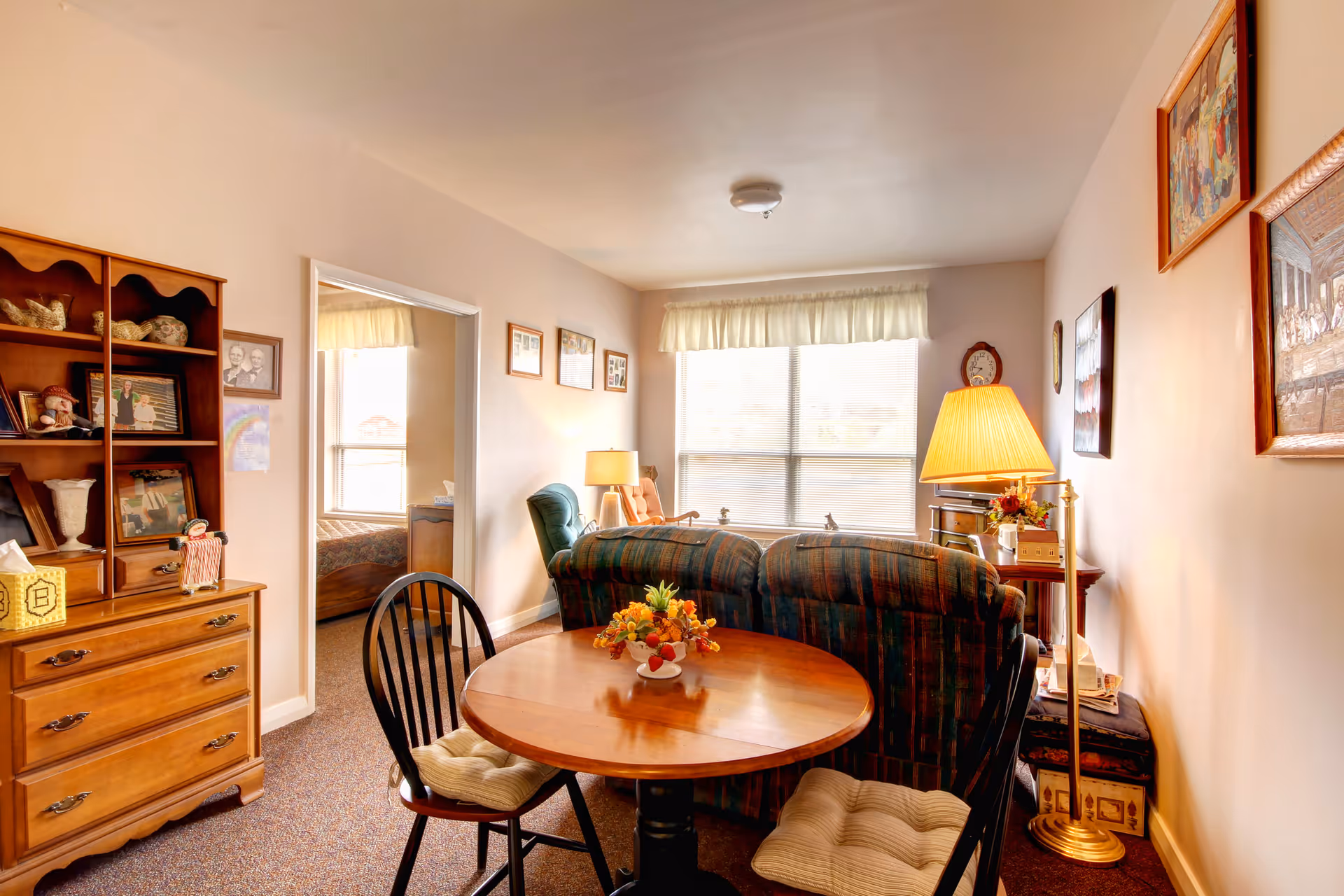 A cozy living area in a senior living facility with a round wooden table and two cushioned chairs in the foreground. Behind the table are two plaid upholstered armchairs facing a large window with blinds and a sheer curtain valance. A floor lamp with a yellow shade stands next to the chairs. On the left side, there is a wooden dresser with shelves displaying framed photos and decorative items. A doorway leads to a bedroom with a bed and window visible. The walls are adorned with framed pictures and artwork.