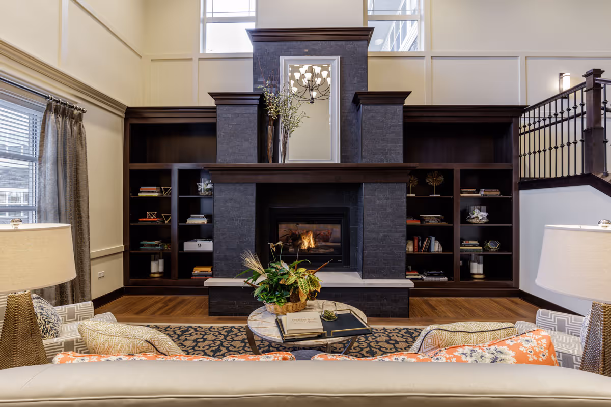 A cozy living room area with a modern fireplace centered between two dark wood built-in bookshelves filled with books and decorative items. Above the fireplace is a large mirror reflecting a chandelier. The room features high ceilings with tall windows, a patterned rug, a round coffee table with a plant and books, and comfortable seating with patterned cushions. Two lamps with white shades flank the seating area.