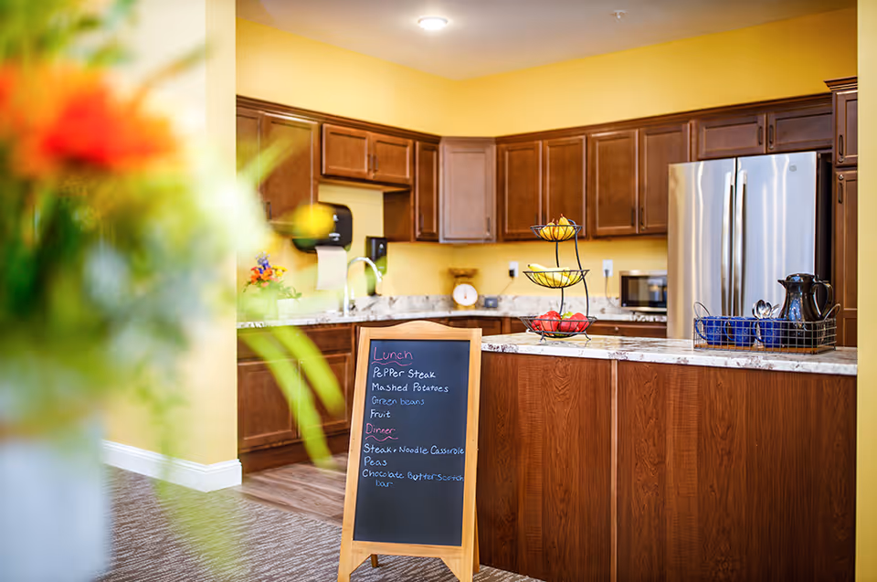Interior view of a kitchen area with wooden cabinets, a stainless steel refrigerator, and a marble countertop island. A small chalkboard on a stand displays the lunch and dinner menu. A tiered fruit basket with bananas and apples is on the countertop. The walls are painted yellow and there is a blurred flower arrangement in the foreground.
