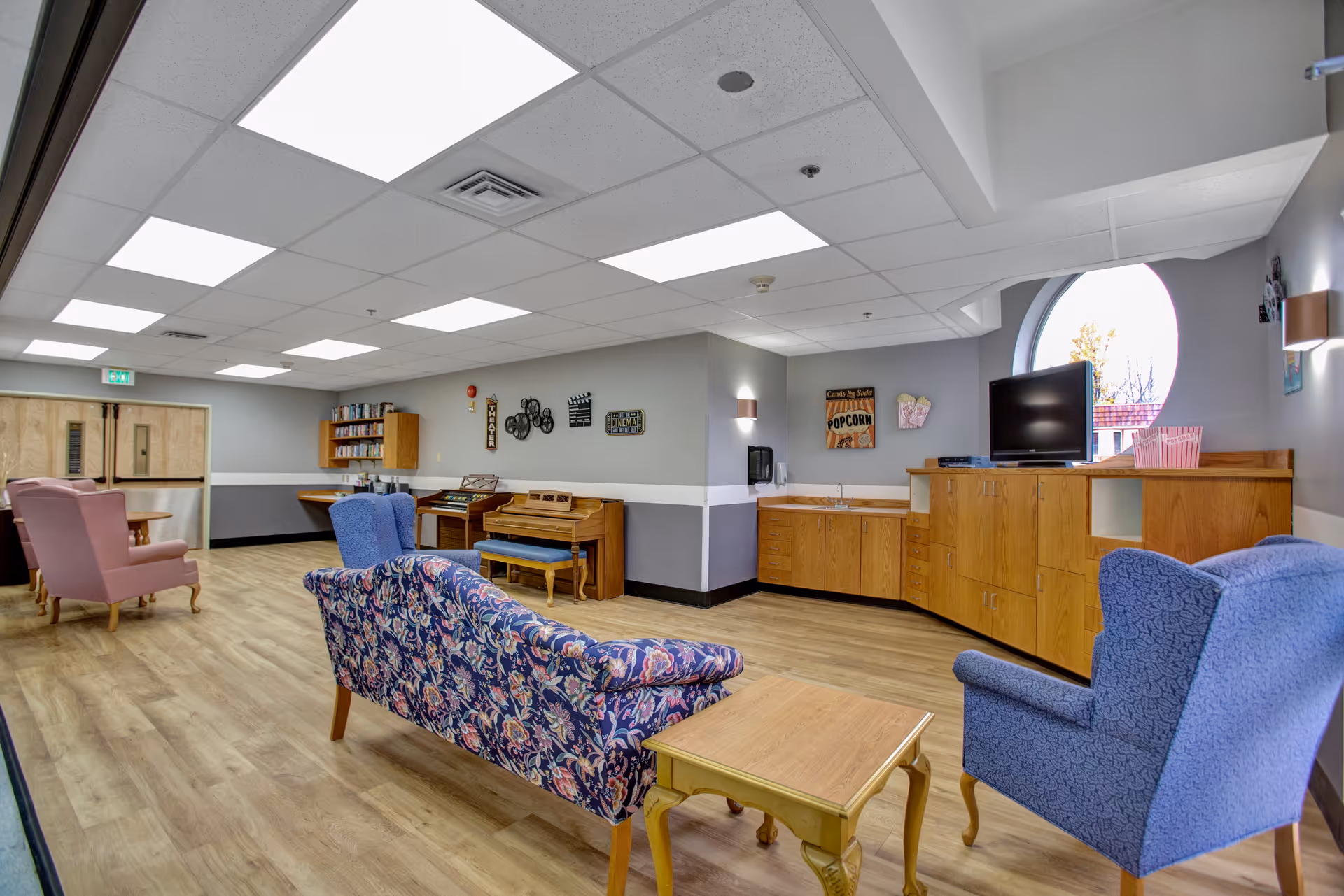 A spacious common area in a senior living facility with wooden flooring, floral and solid-colored upholstered chairs, a wooden coffee table, a piano with a bench, a TV on a wooden cabinet, and wall decorations including a popcorn sign. The room has a large circular window and bright ceiling lights.