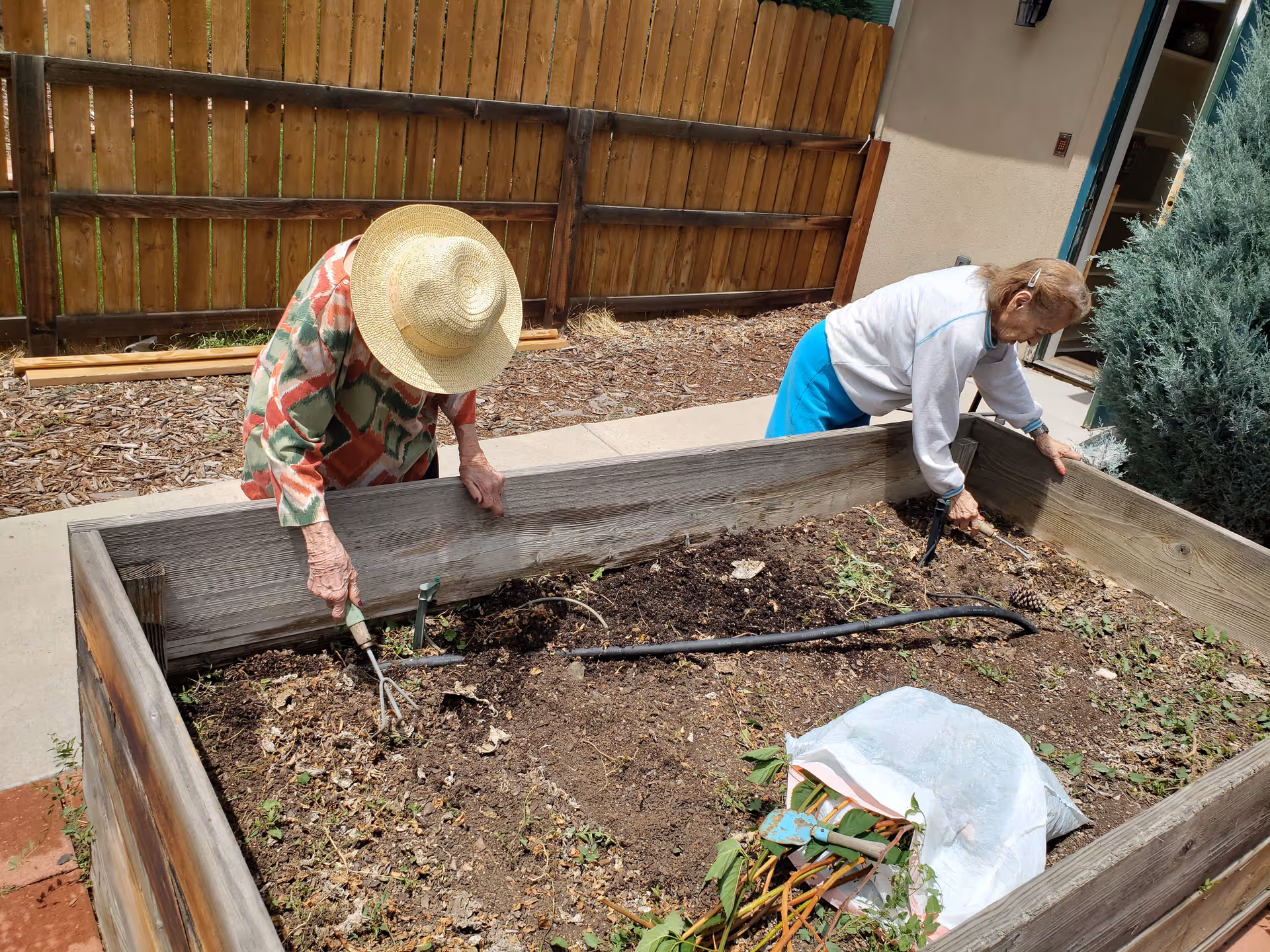 Two elderly women gardening in a raised wooden garden bed outdoors. One woman wears a large straw hat and a colorful patterned shirt, while the other wears a white sweater and blue skirt. They are both using small gardening tools to tend the soil. A wooden fence and some greenery are visible in the background.