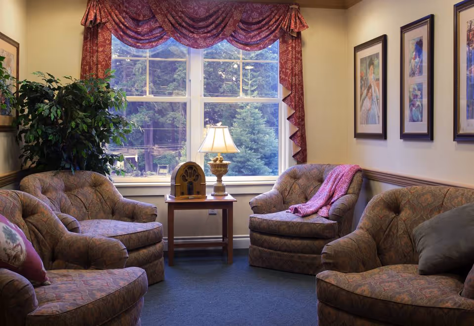 A cozy living room with four patterned armchairs arranged around a wooden side table with a vintage radio and a lamp. The room has a large window with red patterned valances, framed pictures on the wall, and a large green plant in the corner.