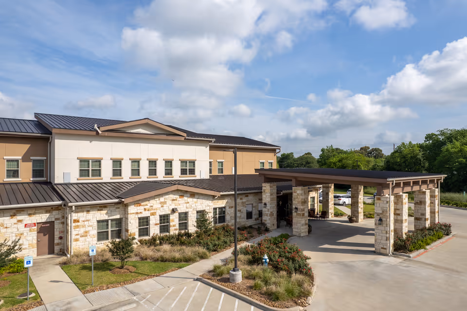 Exterior view of a senior living facility building with a covered entrance supported by stone pillars, surrounded by landscaped greenery and a parking area under a partly cloudy sky.