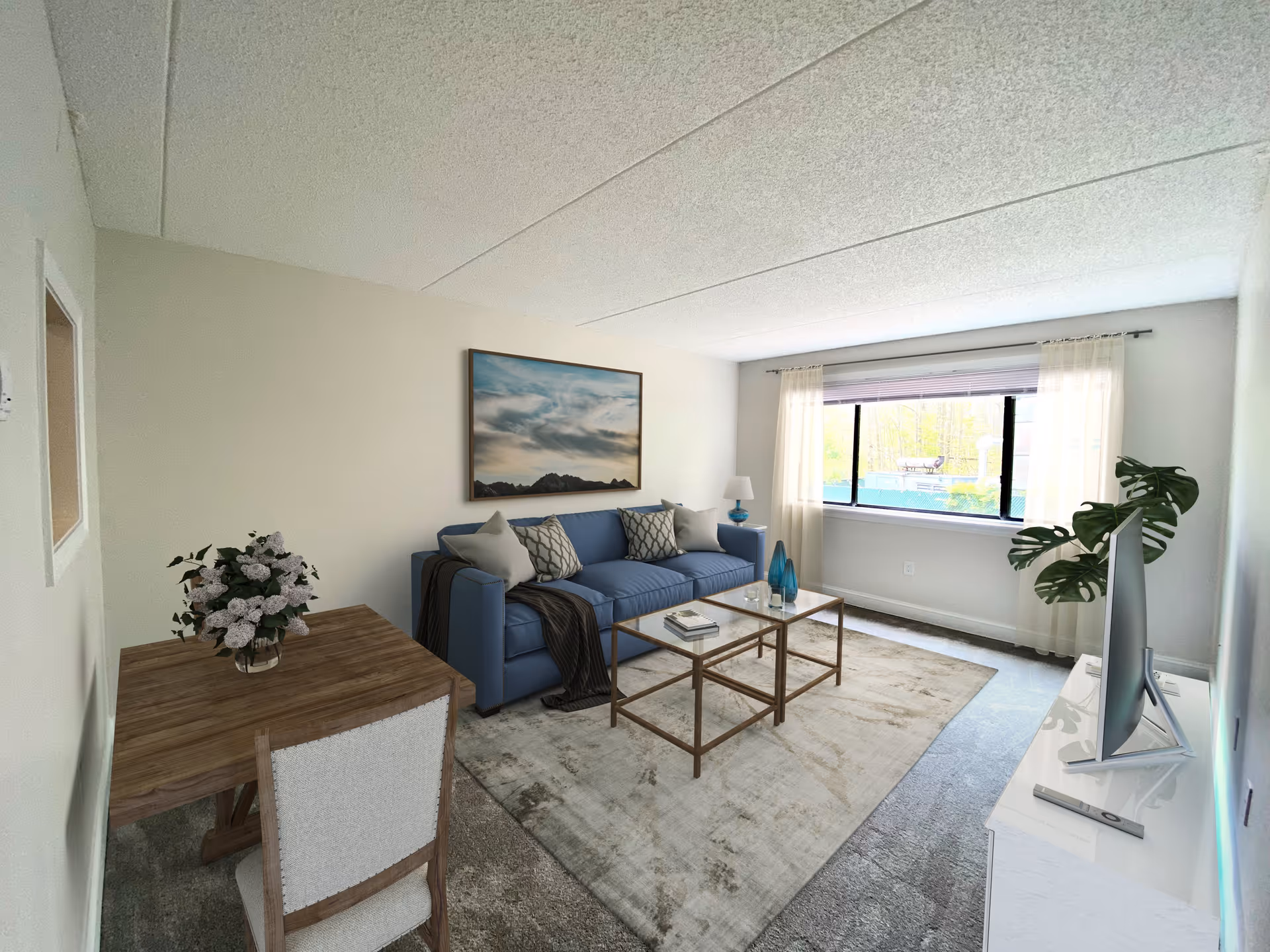 A bright living room with a large window covered by sheer curtains. The room features a blue sofa with patterned and plain cushions, a wooden coffee table with glass top, a wooden dining table with a chair and a vase of flowers, a white TV stand with a flat-screen TV, and a large framed landscape painting on the wall above the sofa. There is a green plant near the TV stand and the floor is covered with a light-colored area rug.