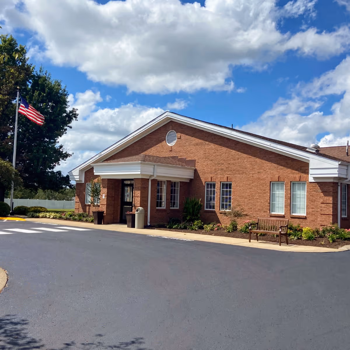 Front entrance of a single-story brick building with a covered portico, American flag, landscaping, and a bench under a blue sky.