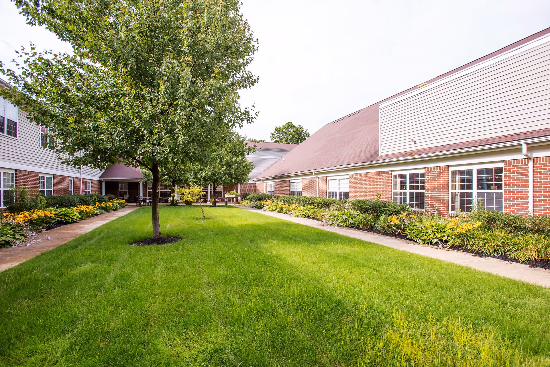 A courtyard area with green grass, trees, and flower beds surrounded by a brick building with white siding and multiple windows under a cloudy sky.