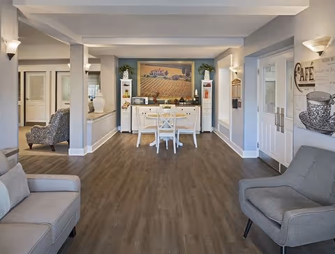 A cozy senior living common area with light gray walls and wood flooring. The room features a white table with two chairs in the center, flanked by two white cabinets with decorative items and plants on top. A large framed painting of a countryside scene hangs on the back wall. There are two comfortable armchairs, one gray and one patterned, positioned on either side of the room. Wall sconces provide soft lighting, and a decorative sign with the word 'CAFE' is visible on the right wall near double doors.