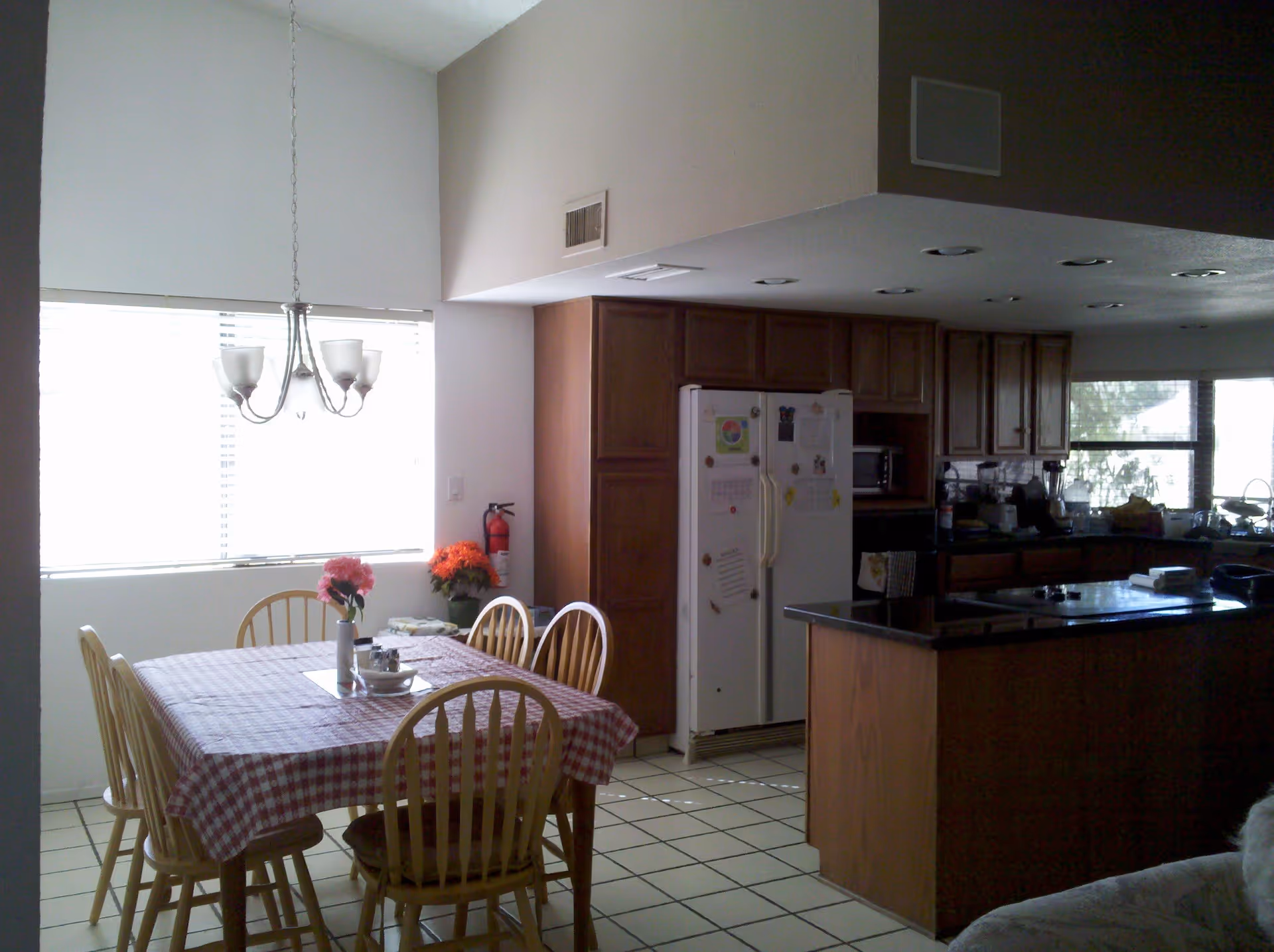 Interior view of a kitchen and dining area with a wooden dining table covered with a red and white checkered tablecloth, surrounded by six wooden chairs. A vase with flowers is placed on the table. The kitchen has wooden cabinets, a white refrigerator with magnets and notes, a black countertop island, and various kitchen appliances. A window with blinds lets in natural light, and a chandelier hangs above the dining table.