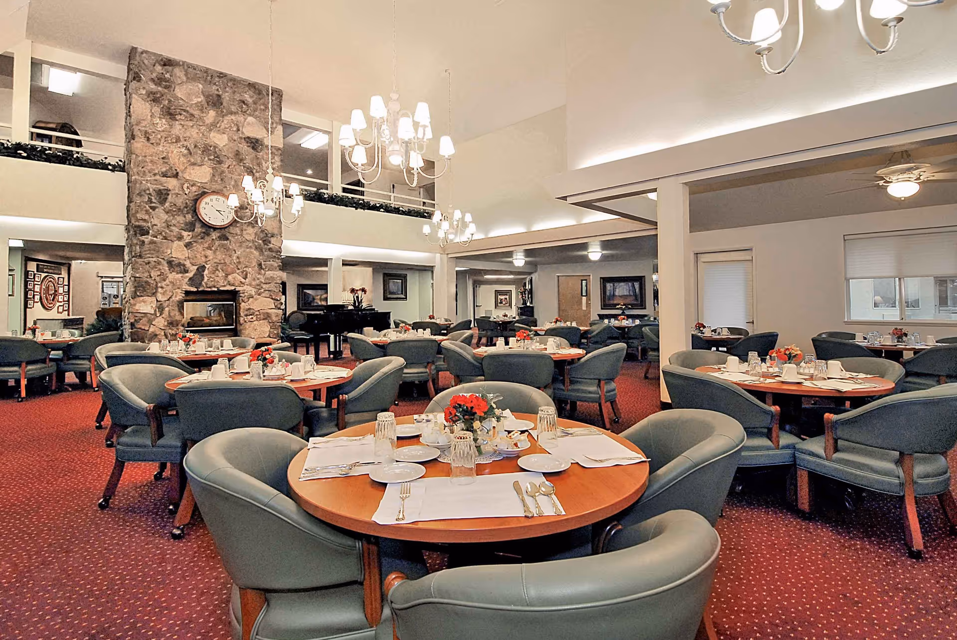 A spacious dining room with round wooden tables set with white plates, glasses, silverware, and small flower arrangements. The room features green upholstered chairs, a red carpet, a large stone fireplace with a clock above it, chandeliers hanging from the ceiling, and a piano in the background.