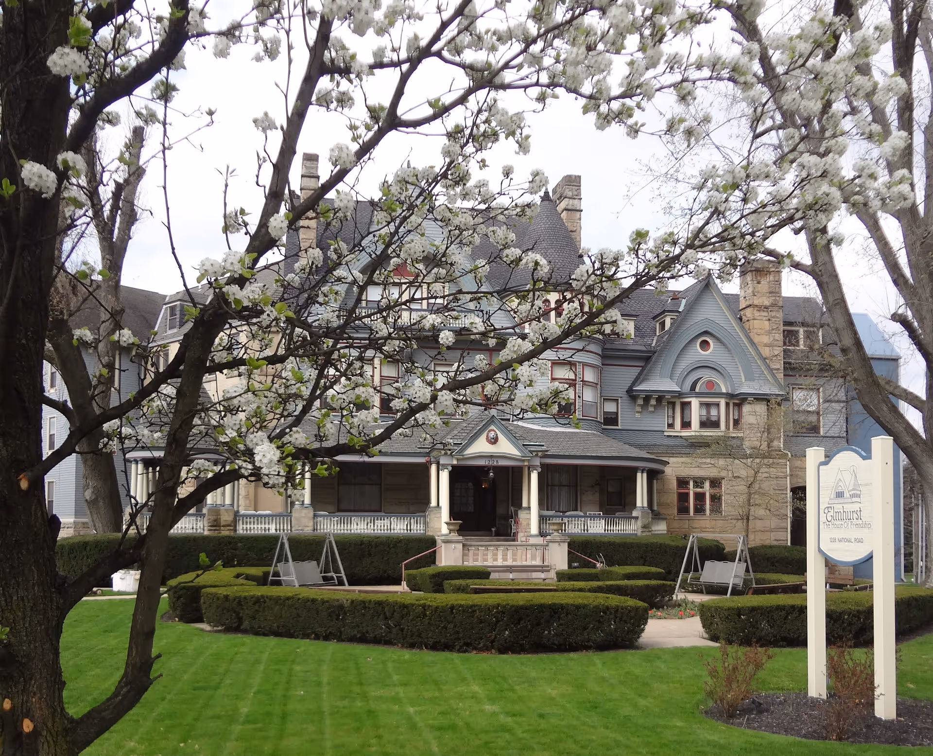A large, historic-style building with a steep roof and multiple chimneys, partially obscured by a tree with white blossoms in the foreground. The building is surrounded by neatly trimmed hedges and a well-maintained lawn. In front of the building, there is a sign that reads 'Elmhurst - The House Of Friendship'.