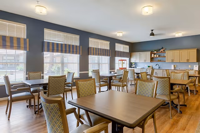 A bright dining room with multiple wooden tables and cushioned chairs arranged neatly. Large windows with striped valances allow natural light to fill the room. The back wall features a kitchenette area with cabinets, a countertop, and shelves holding decorative items.