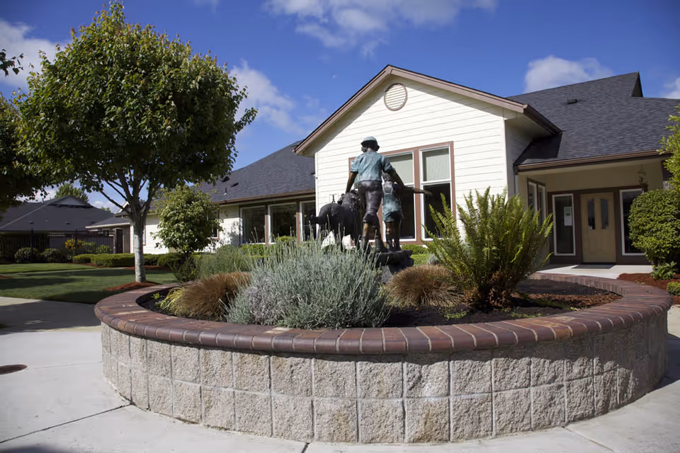 Front exterior of a single-story building with a circular raised planter, statue, and landscaped grounds.