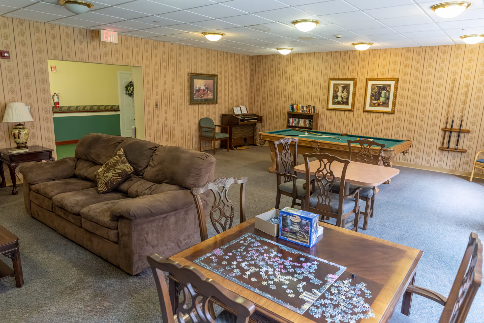 Community common room with a brown sofa, tables and chairs with a jigsaw puzzle, and a pool table against patterned wallpaper.