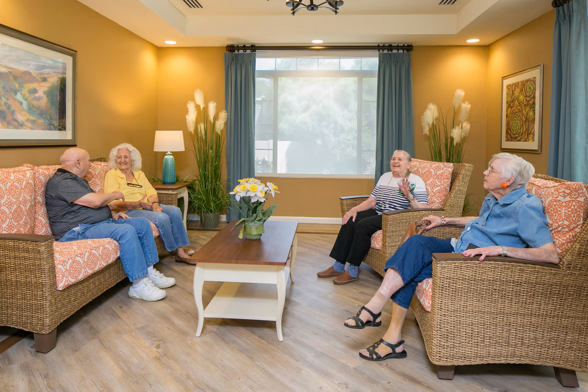 A cozy living room in Orchard Pointe at Arrowhead with four elderly people sitting on wicker furniture with orange patterned cushions, engaged in conversation. The room features a wooden coffee table with a flower arrangement, a large window with blue curtains, warm yellow walls, framed artwork, and decorative plants.