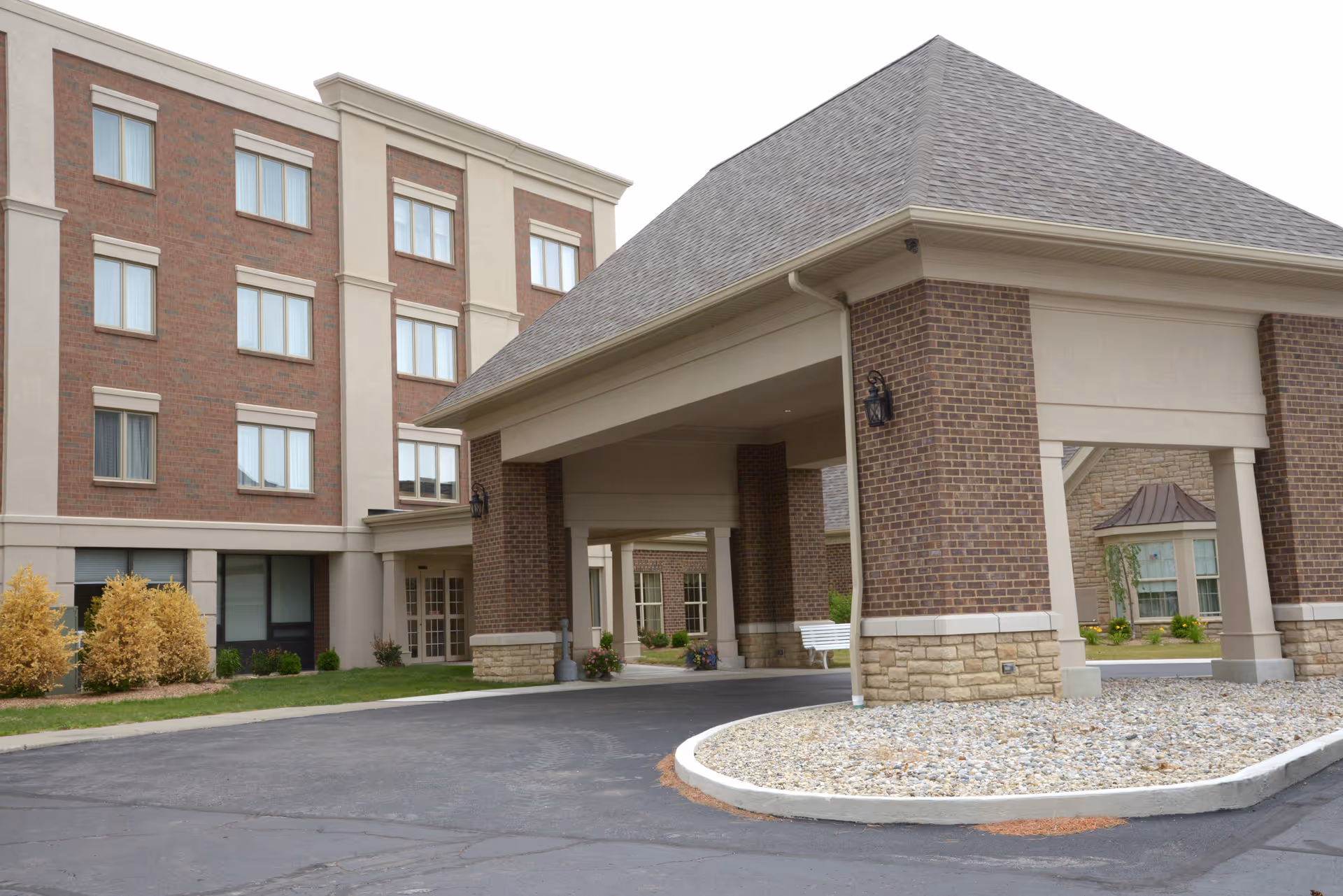 Exterior view of a multi-story senior living facility building with a covered entrance driveway. The building features brick and beige siding with multiple windows. There are small landscaped areas with bushes and a paved driveway in front.
