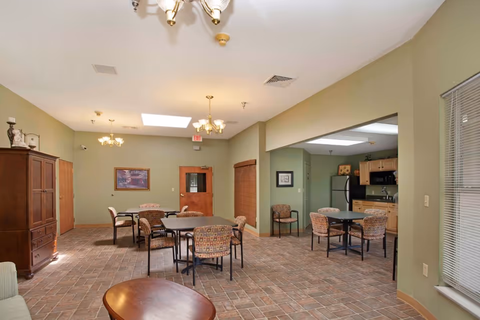 A spacious common area with multiple tables and chairs arranged for seating. The room has green walls, tile flooring, and ceiling lights. There is a kitchenette with wooden cabinets, a refrigerator, and a microwave visible in the background. A wooden cabinet and a framed picture are on the walls, and a window with blinds is on the right side.
