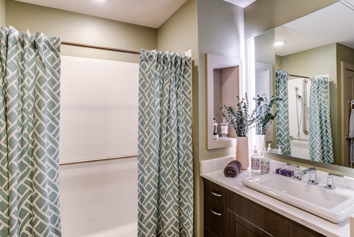 A bathroom with a shower area enclosed by a patterned blue and white shower curtain. There is a large mirror above a white sink with a modern faucet, set in a countertop with dark wooden cabinets below. On the countertop, there is a rolled dark towel, a vase with green foliage, and various soap dispensers. The walls are painted light green.