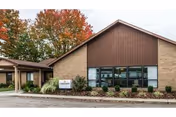 Exterior view of Belmont Healthcare Center, a single-story building with beige brick walls and a brown gabled roof. There are large windows on the front, landscaping with shrubs and plants, and trees with autumn-colored leaves in the background. A sign near the entrance displays the facility's name.