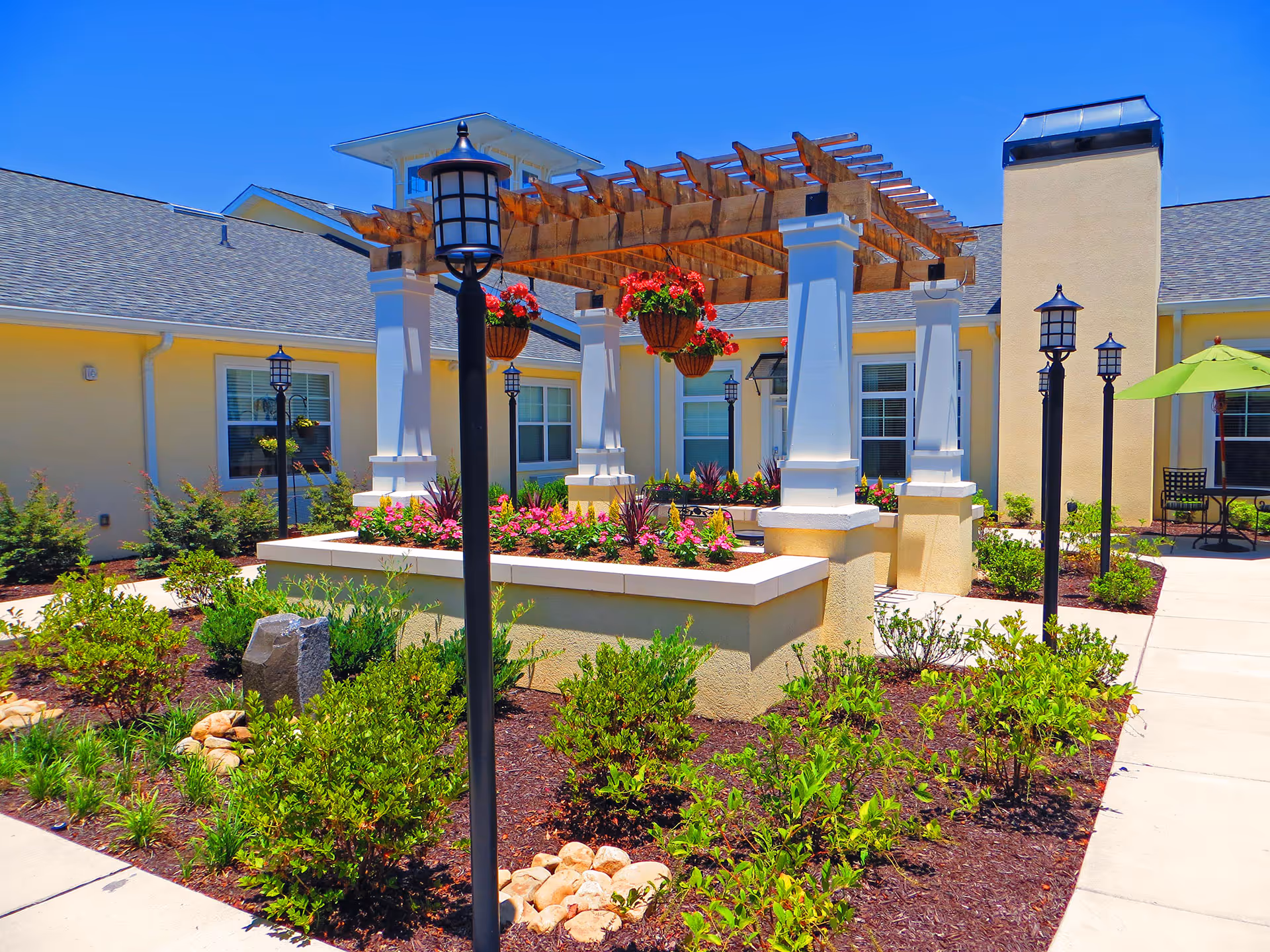 Outdoor courtyard area at Barclay House of Augusta featuring a pergola with hanging flower baskets, surrounded by landscaped garden beds with various green plants and flowers. The courtyard is bordered by a light yellow building with windows and outdoor seating under a green umbrella. Several black lamp posts line the walkway.
