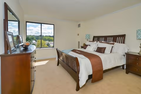 Sunny bedroom with a wooden bed, matching dresser, bedside tables, and a large window showing a tree-lined view.