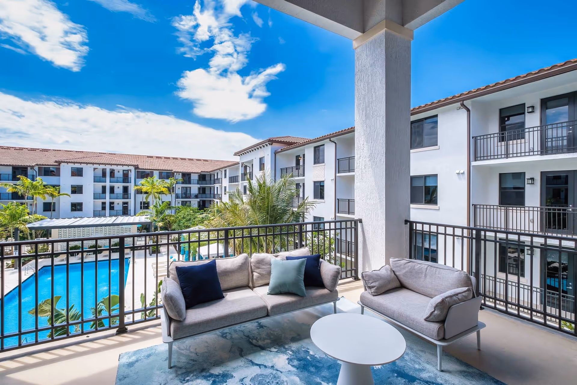 Covered balcony with cushioned seating and a small table overlooking a swimming pool and multi-story residential buildings under a blue sky.