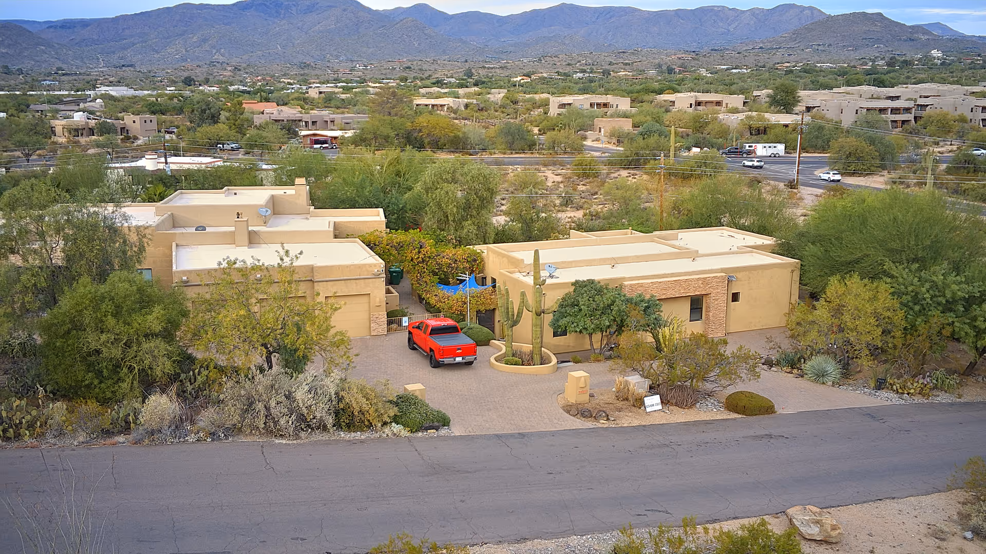 Aerial view of a single-story beige stucco building with a red pickup truck in the driveway, desert landscaping and mountains in the background.